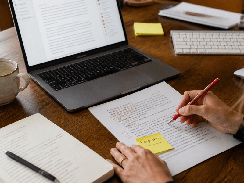 A person marking up a printed draft beside a Mac, with a yellow sticky note reading Keep this on the strongest passage