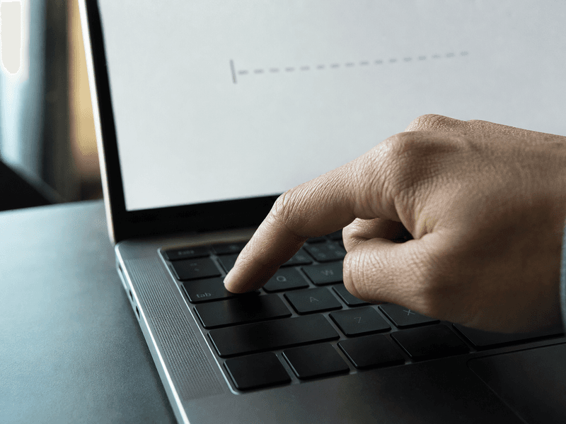 A close-up of a hand pressing the Tab key on a MacBook while a faint autocomplete suggestion appears on screen
